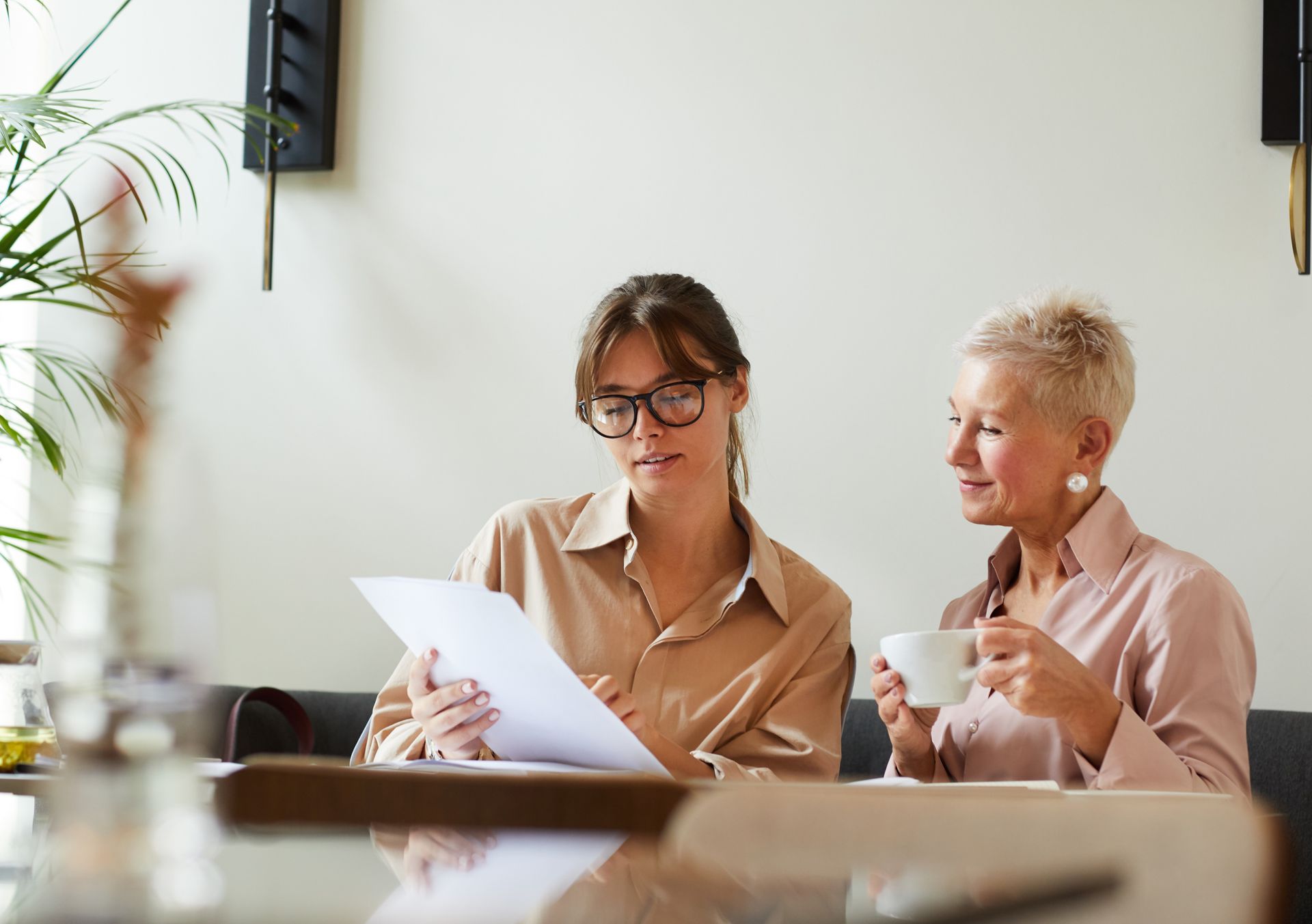 Two women in a cafe reviewing documents; one holds papers, the other sips coffee.