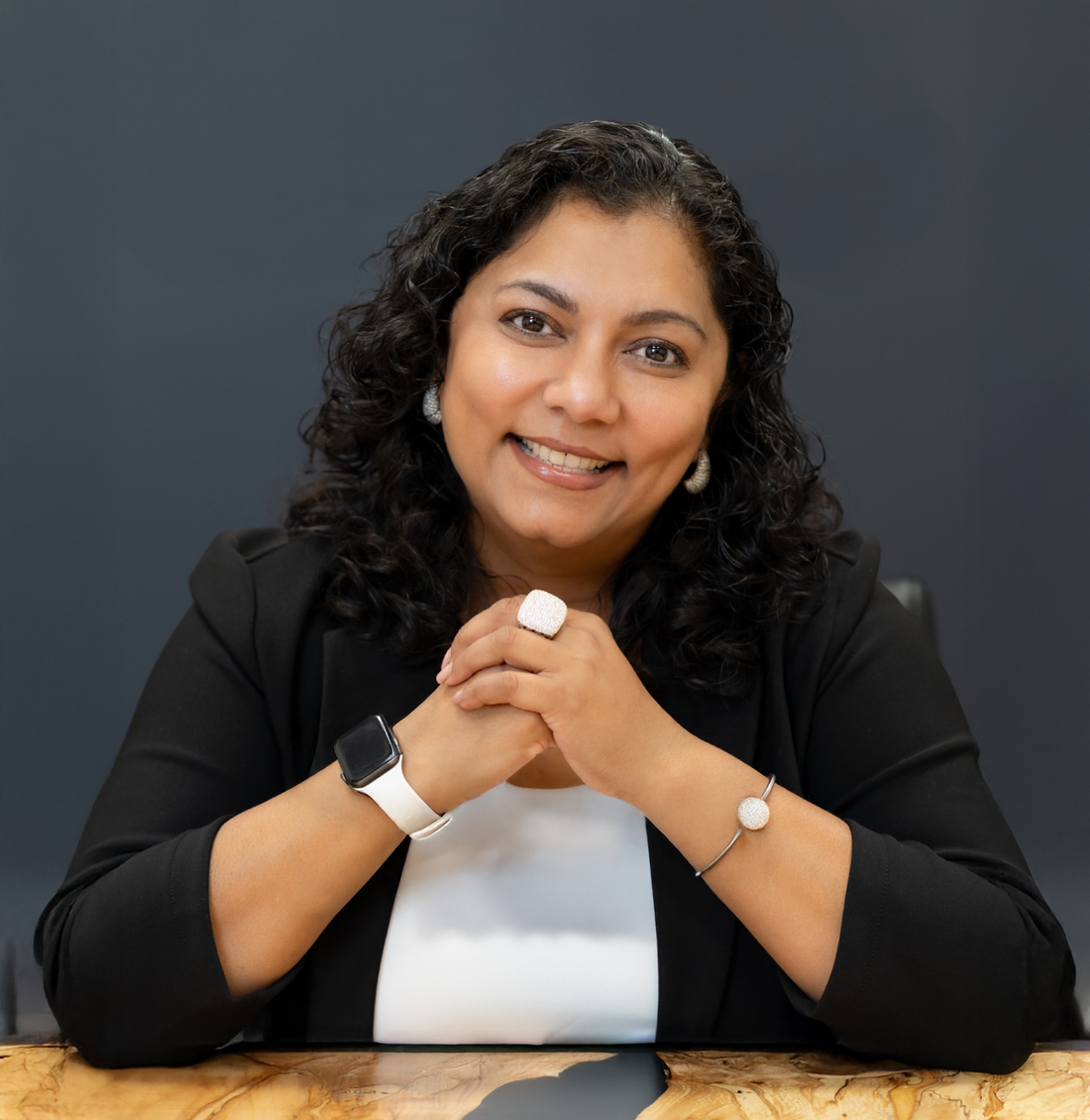 Woman with curly black hair, wearing a black blazer, smiles with hands clasped on a dark desk; grey background.
