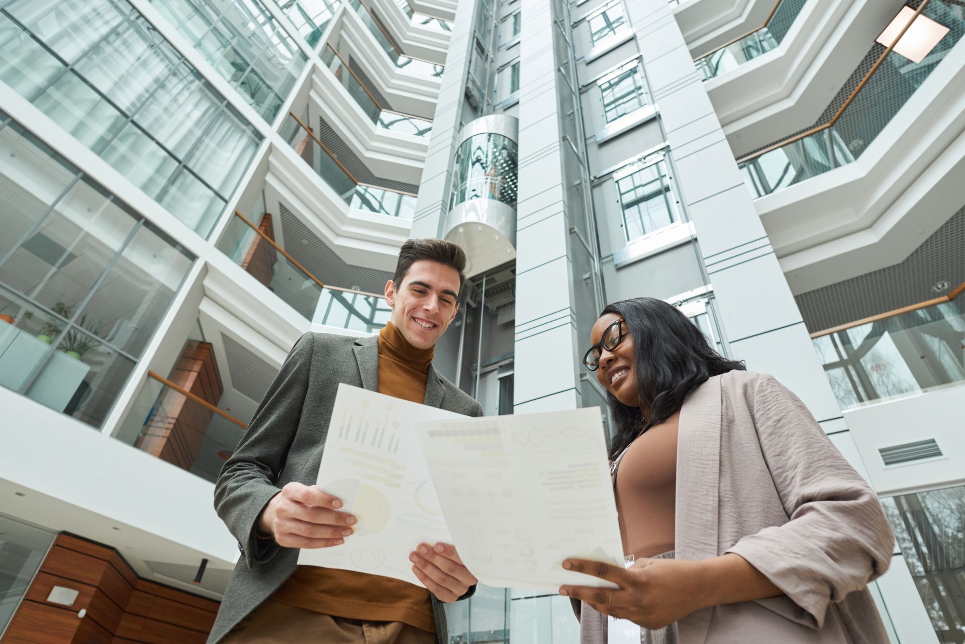 Two businesspeople in a modern building, smiling while reviewing papers. A tall, glass-walled atrium is in the background.