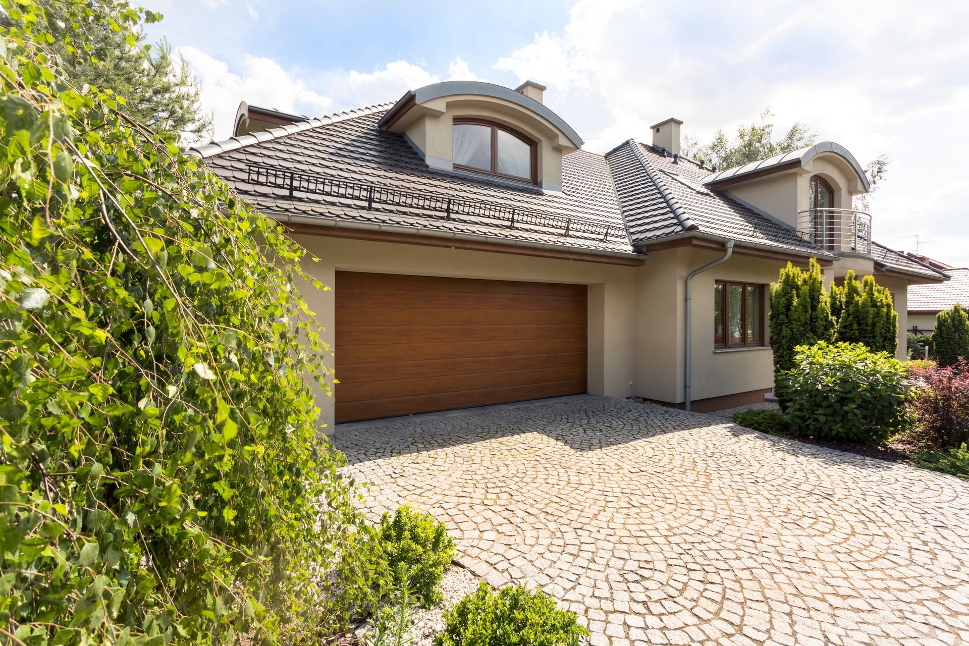Tan house with a brown garage door and cobblestone driveway. Lush green bushes and a blue sky are visible.