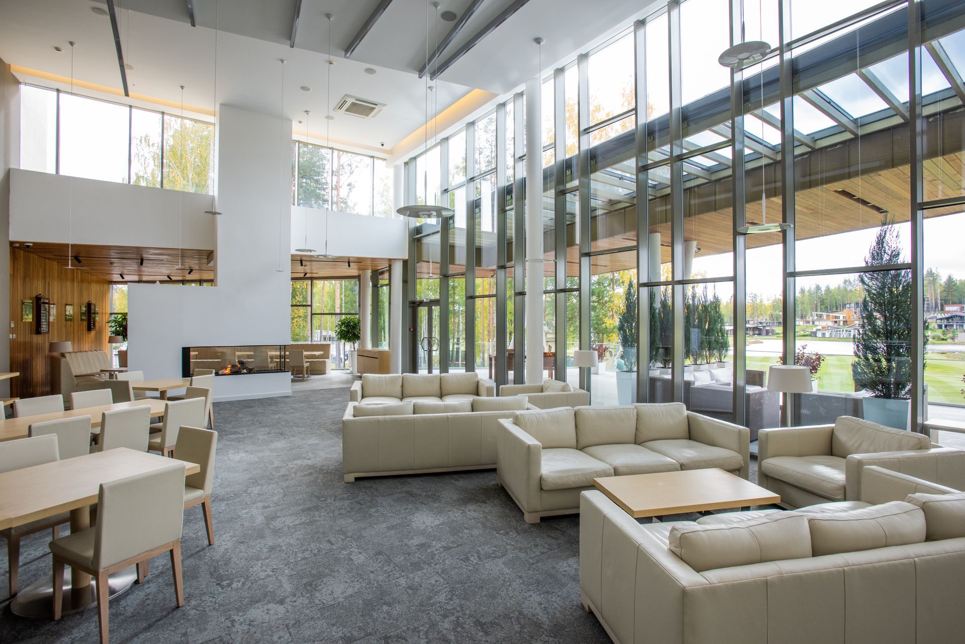 A modern, light-filled lobby with seating areas, tables, and large windows overlooking a grassy area. Beige couches, tables, and chairs populate the room.