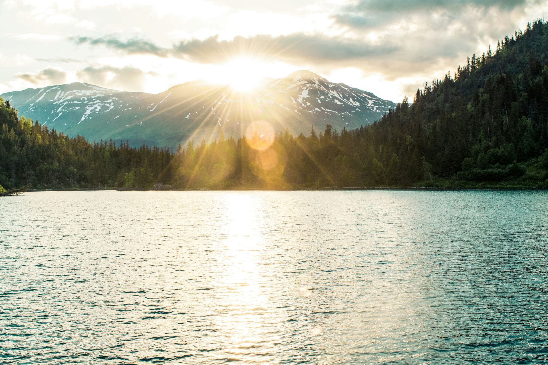 The sun is shining through the clouds over a lake surrounded by mountains.