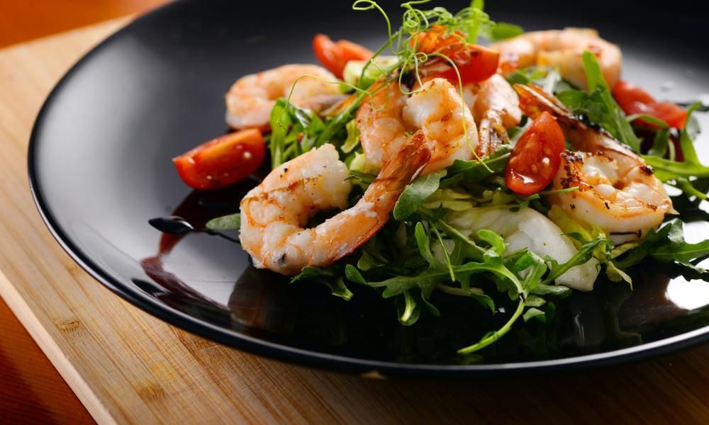 A Black Plate Topped With Shrimp and Tomatoes on a Wooden Table — Noosa Junction Seafood Market in Noosa Heads, QLD