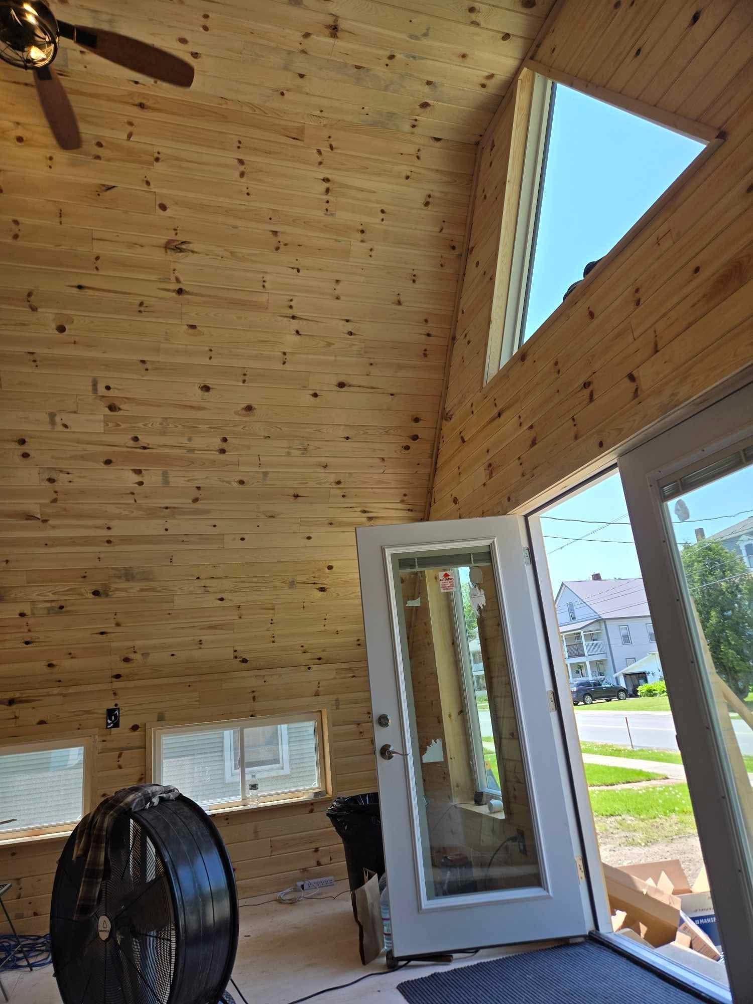 Interior of a room with wooden walls, a large triangle window, and an open door, with a fan and ceiling fan visible.