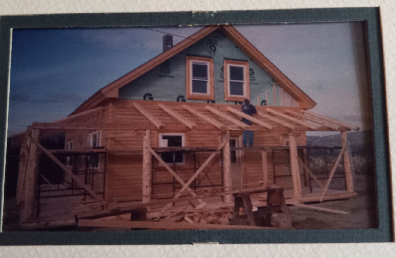 A wooden house under construction; a person on the roof, blue wrap, clear sky.