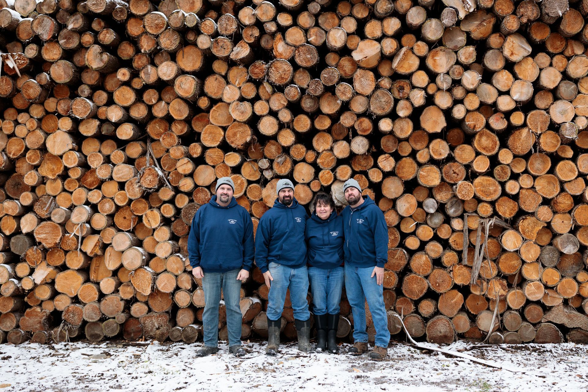 Four people in blue stand before a large pile of cut logs, snow on the ground.