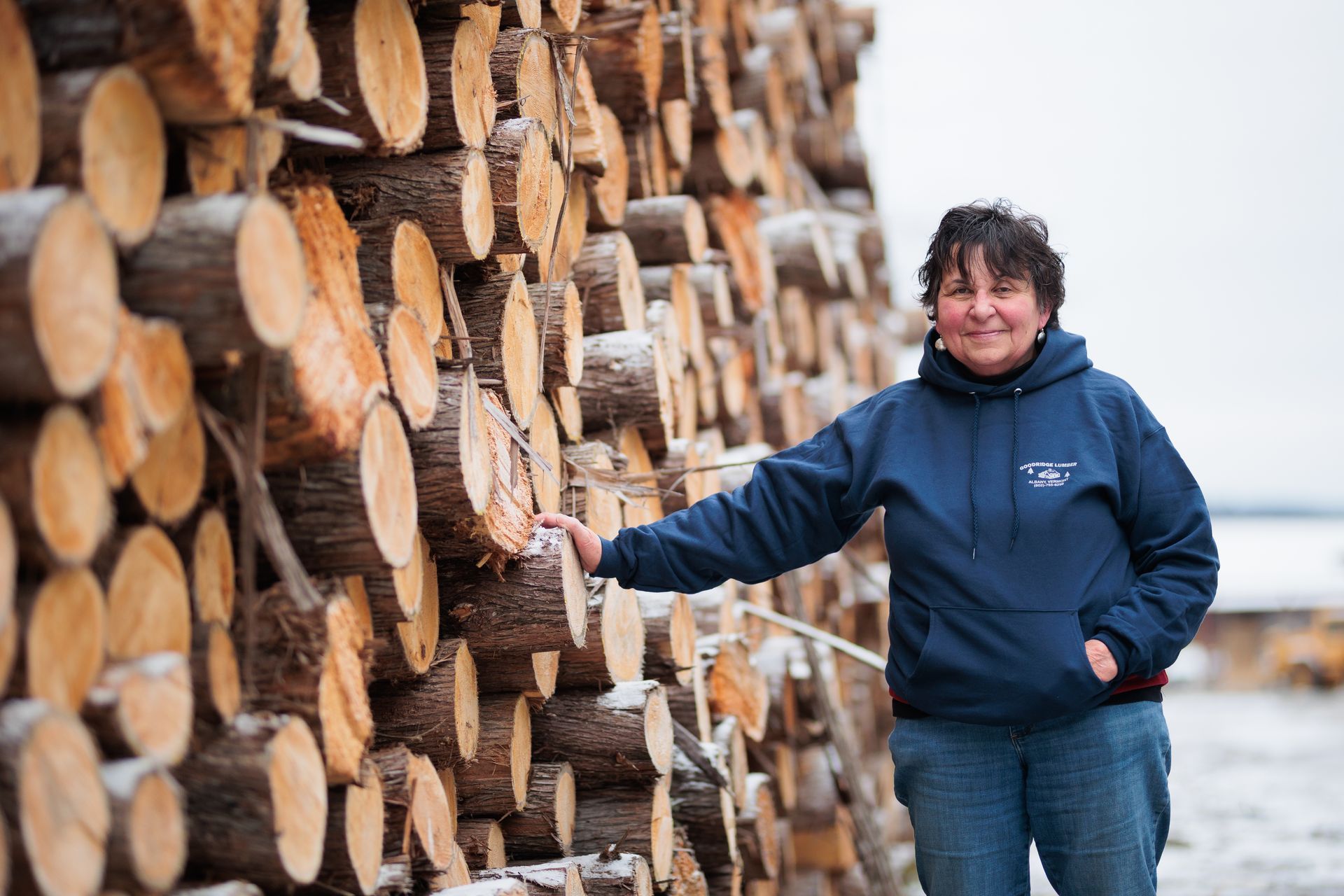 Woman in blue hoodie beside a large stack of freshly cut logs. Outdoor, winter setting.