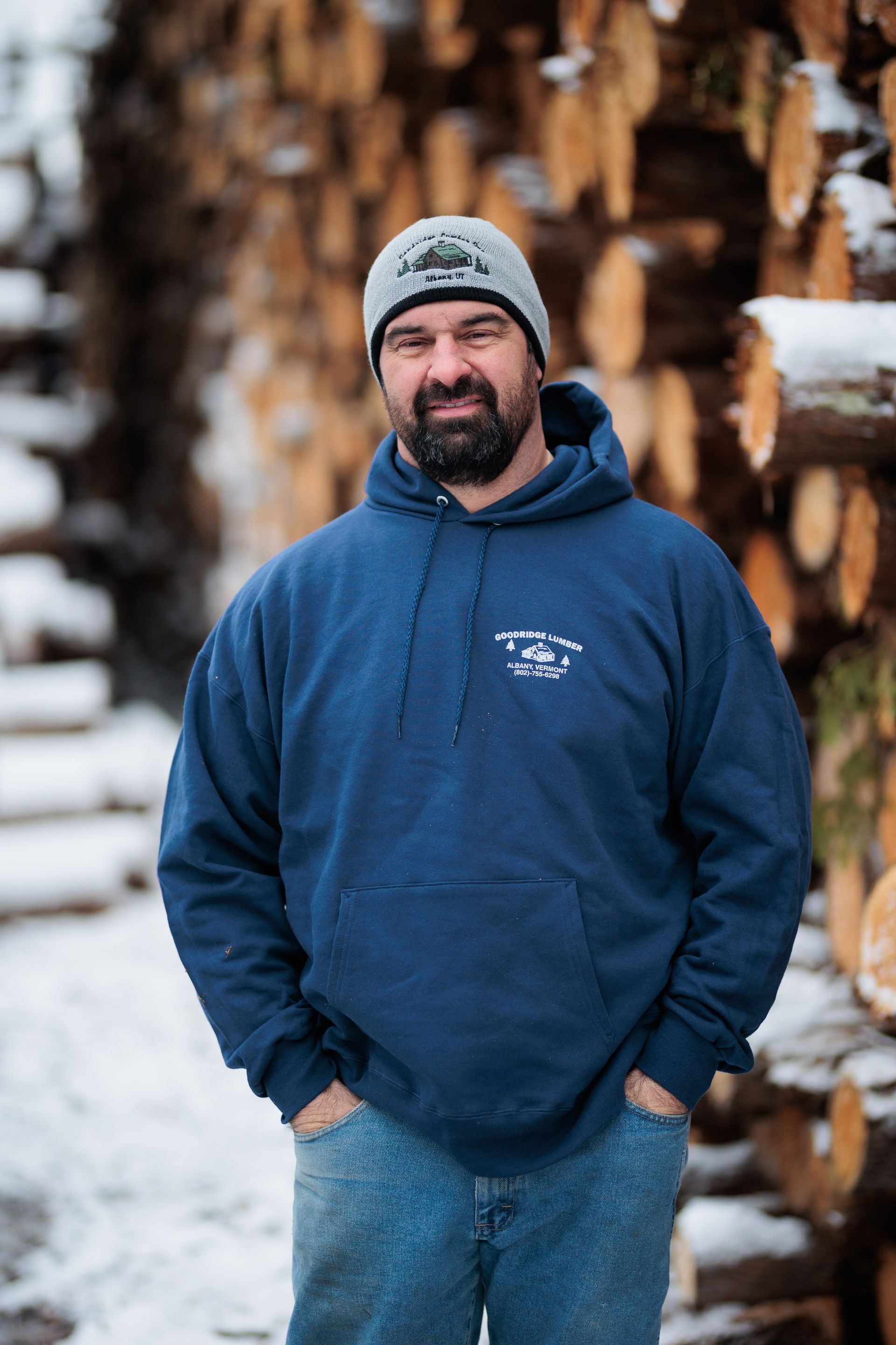 Man in blue hoodie and beanie stands in front of a stack of logs, hands in pockets. Snowy setting.