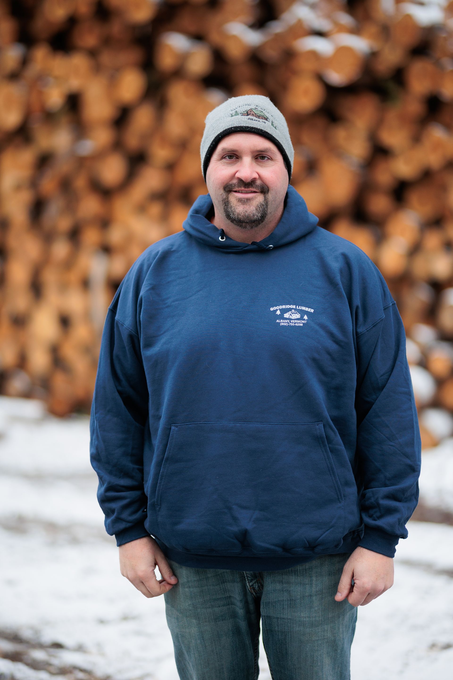 Man in blue hoodie and beanie, standing in front of a pile of logs.