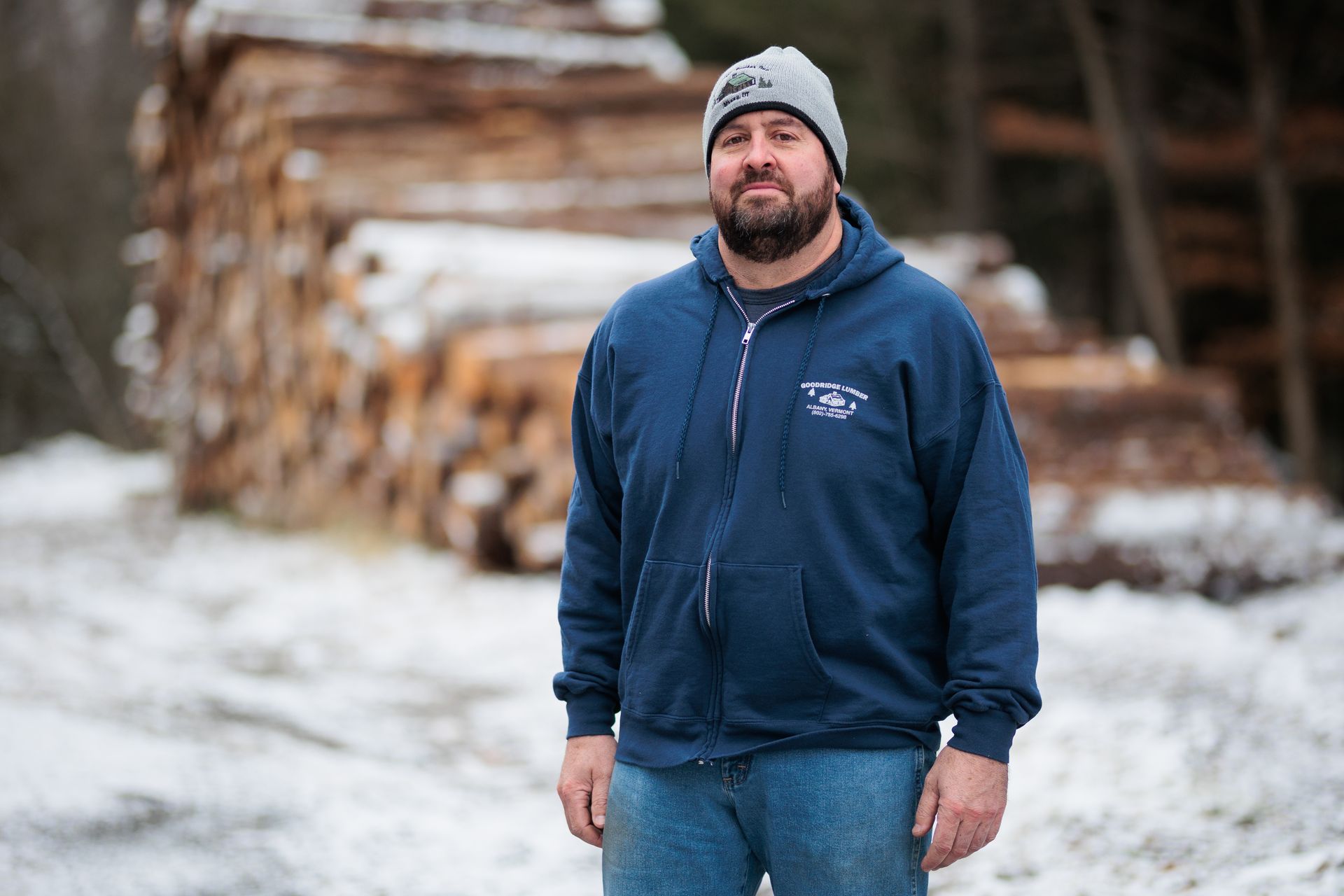 Man in blue hoodie and beanie stands in front of stacked logs in a snowy area.
