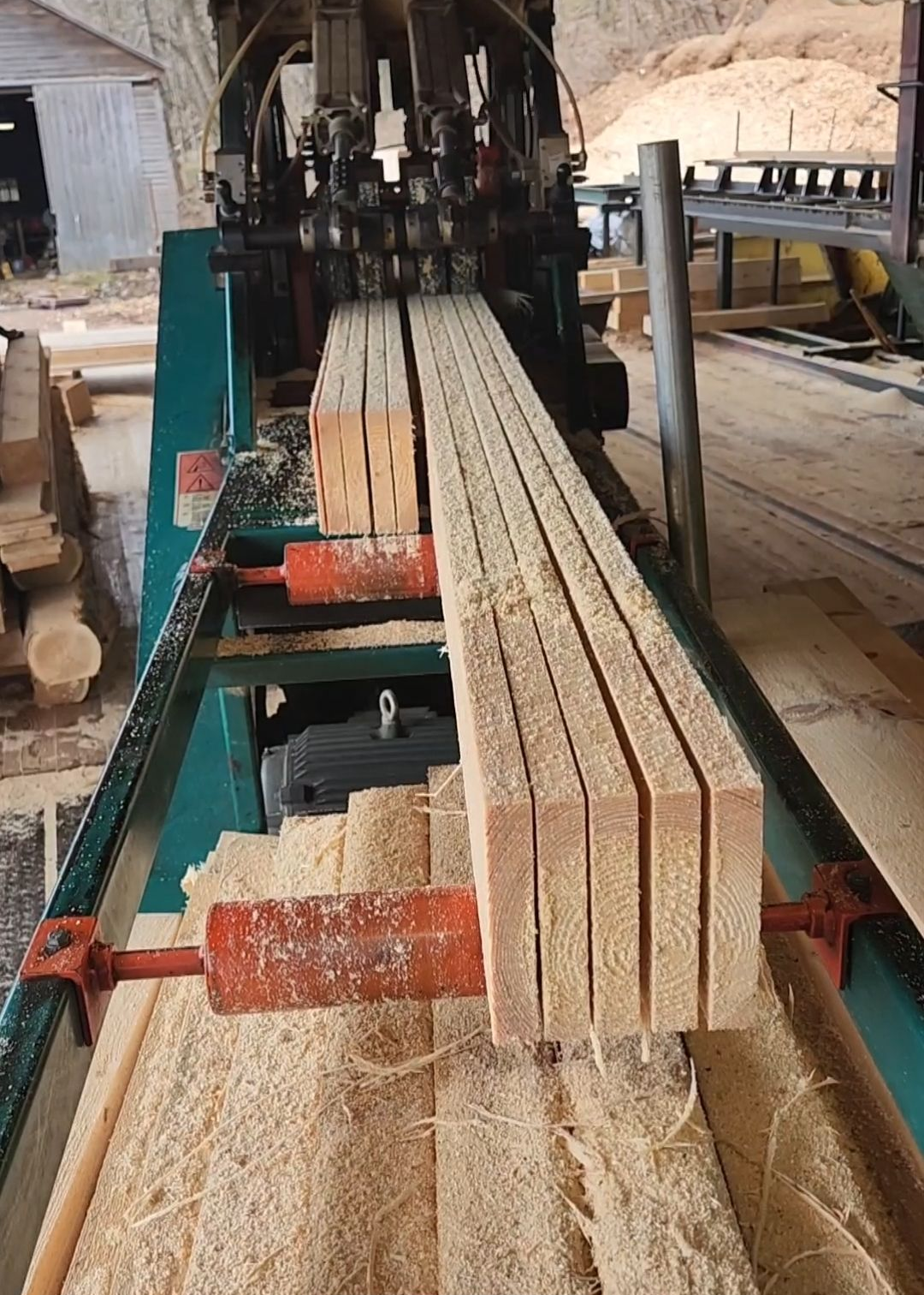 Lumber mill machine cutting freshly sawn wood into uniform boards. Sawdust surrounds the conveyor.
