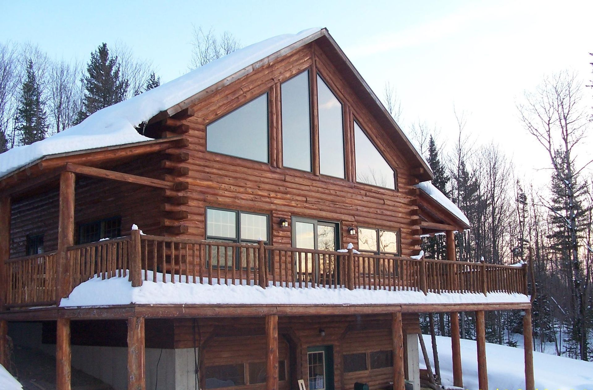Log cabin with snow-covered roof and balcony, tall windows, and a snowy landscape.