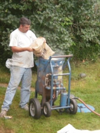 Man pouring material from a bag into a blue sandblaster in a grassy yard.