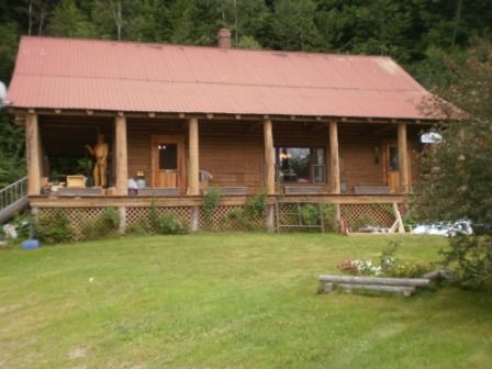 Wooden cabin with a red roof, a wraparound porch, and a green lawn.