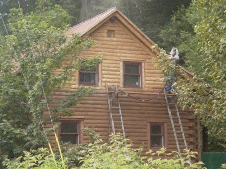 Log cabin house with two ladders. Person on roof. Surrounded by trees.