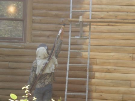 Person wearing protective gear painting a log cabin from a ladder.