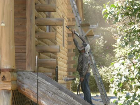 Man spraying log cabin from a ladder. Dust visible. Outdoors in VT.