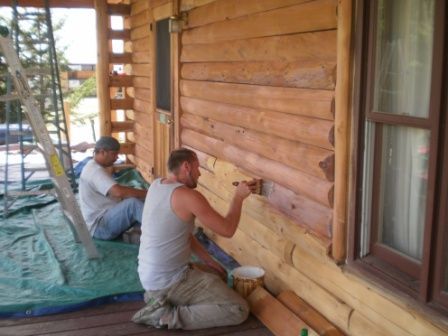 Two men staining the wooden exterior of a log cabin, one painting, the other seated, with ladder and tarp.