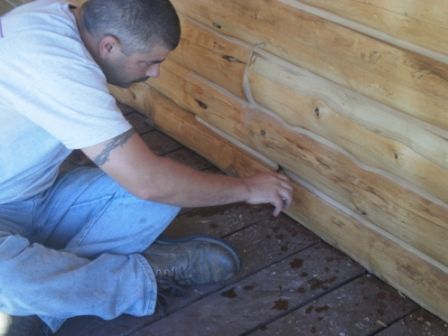 Man kneeling, applying caulk to the logs of a wooden cabin, outside on a wooden deck.