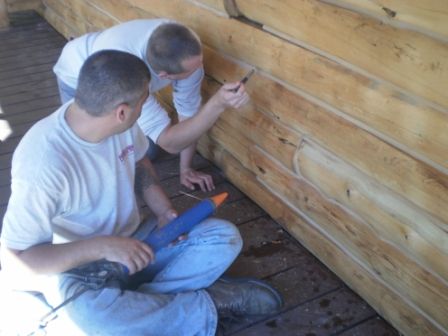 Two men caulking a wooden log house, one seated with a caulk gun, the other pointing.