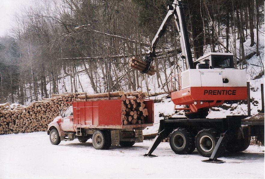 A red Prentice log loader loads a truck with logs in a snowy forest setting.