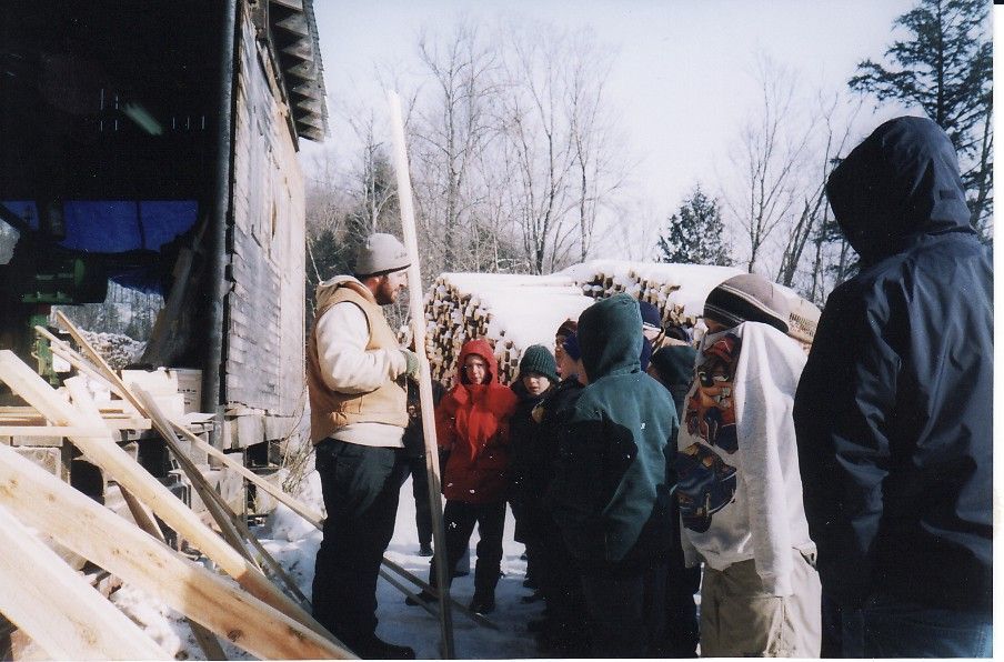 A man speaks to a group outdoors on a snowy day near a wooden structure.