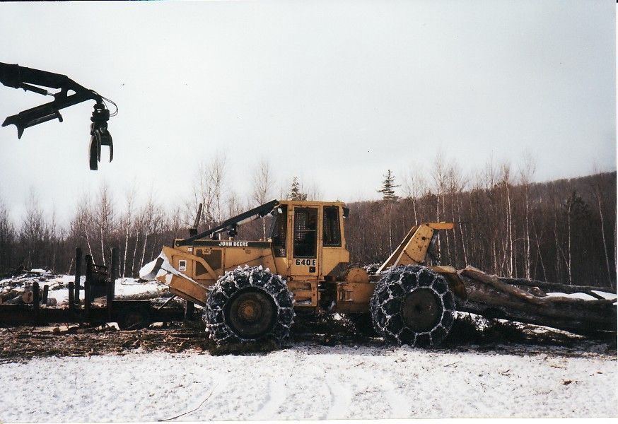 Yellow logging machine with tire chains, hauling logs in a snowy forest.