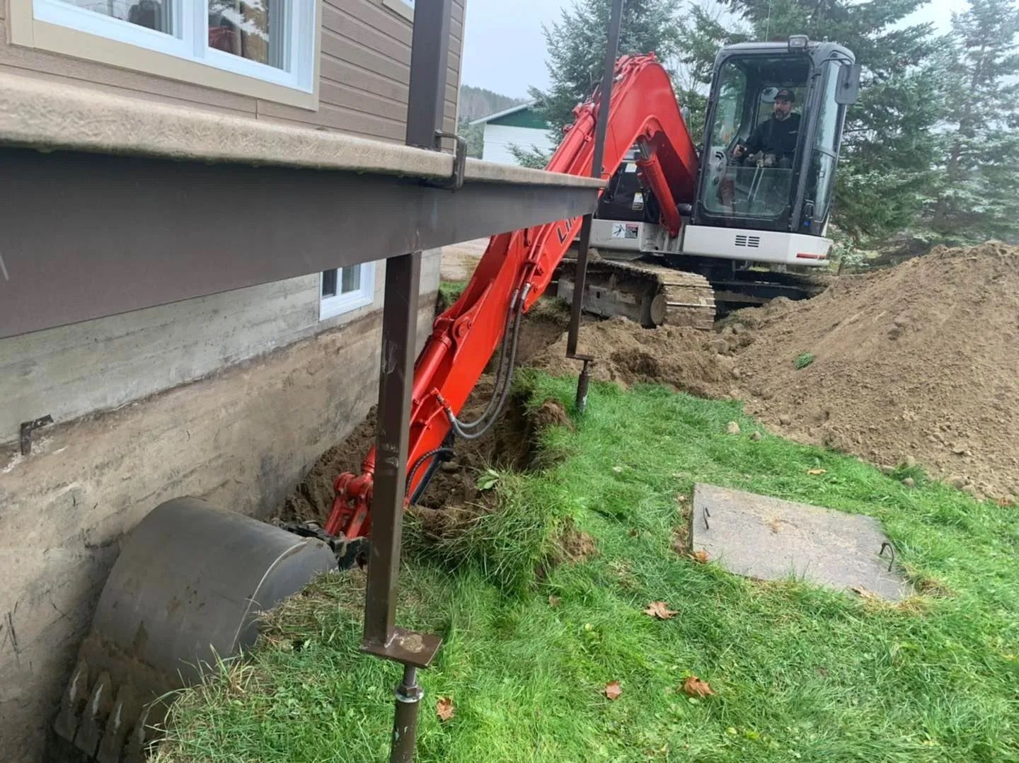 Une pelleteuse orange creuse une tranchée le long des fondations d'une maison, à côté d'un tas de terre.