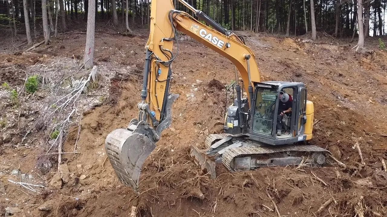 Une pelle mécanique Case jaune creuse un talus de terre dans une zone boisée.
