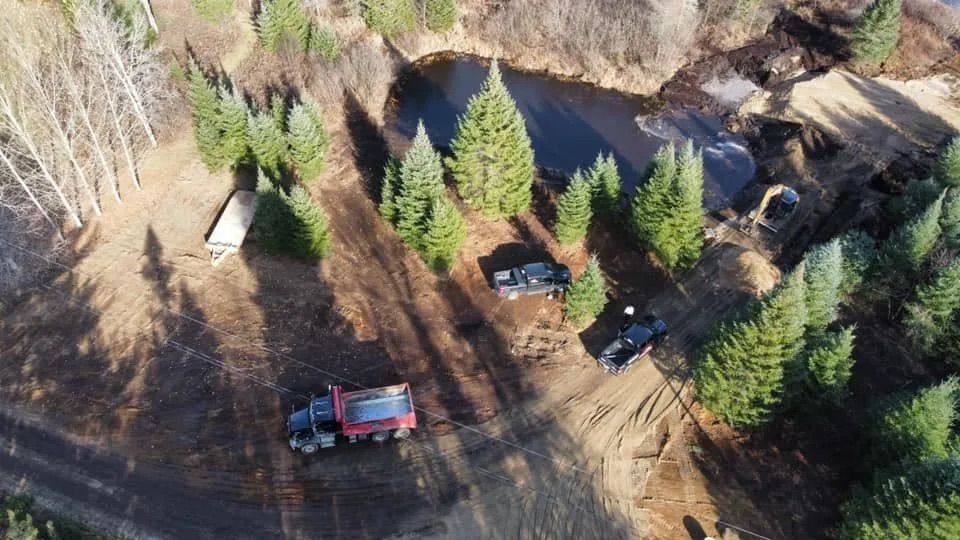 Vue aérienne d'une clairière avec un camion-benne et deux camionnettes garés près d'une rangée d'arbres et d'un étang.