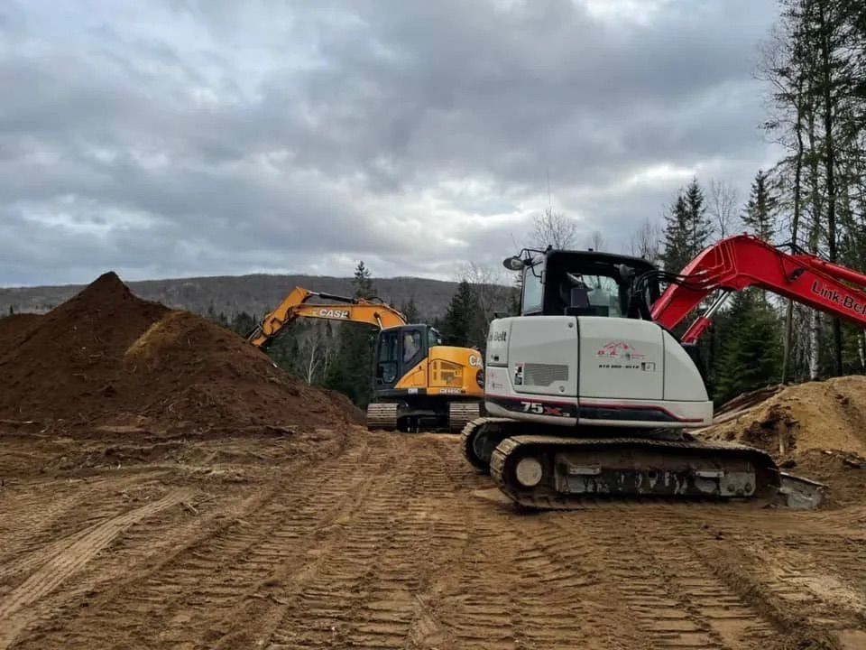 Deux excavatrices, une blanche et une jaune, travaillent sur un chantier de construction en terre, avec des collines et des arbres en arrière-plan.