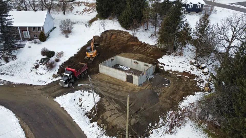 Une vue aérienne montre une excavatrice et un camion-benne à l'œuvre sur un chantier de construction avec des fondations en béton sur un sol enneigé.