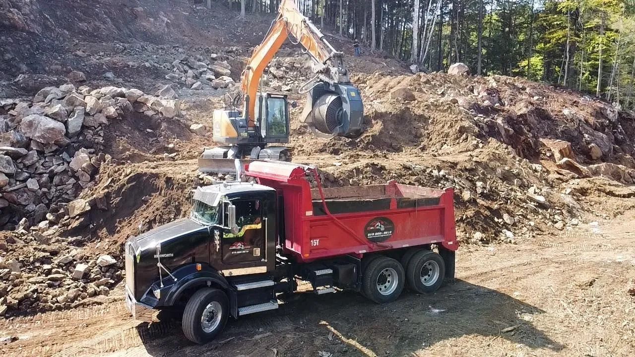 Une excavatrice charge de la terre dans un camion-benne rouge stationné sur un chantier rocailleux et boisé.