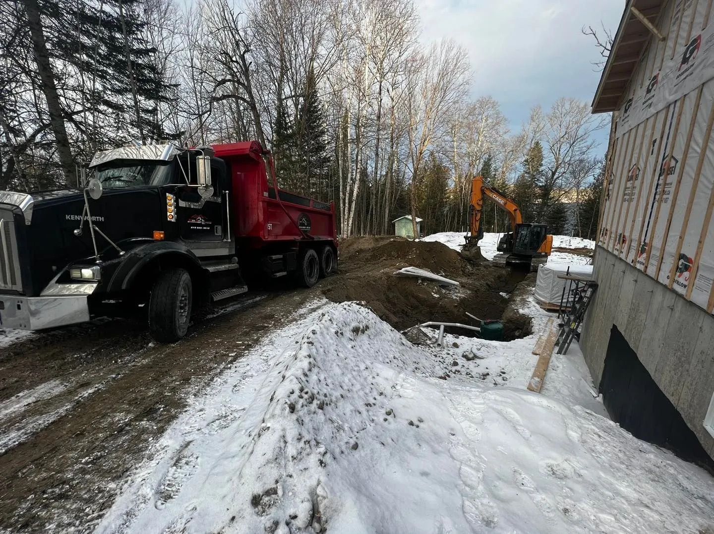 Un camion-benne rouge et une excavatrice effectuent des travaux d'excavation à côté d'un bâtiment en construction sur un chantier enneigé.