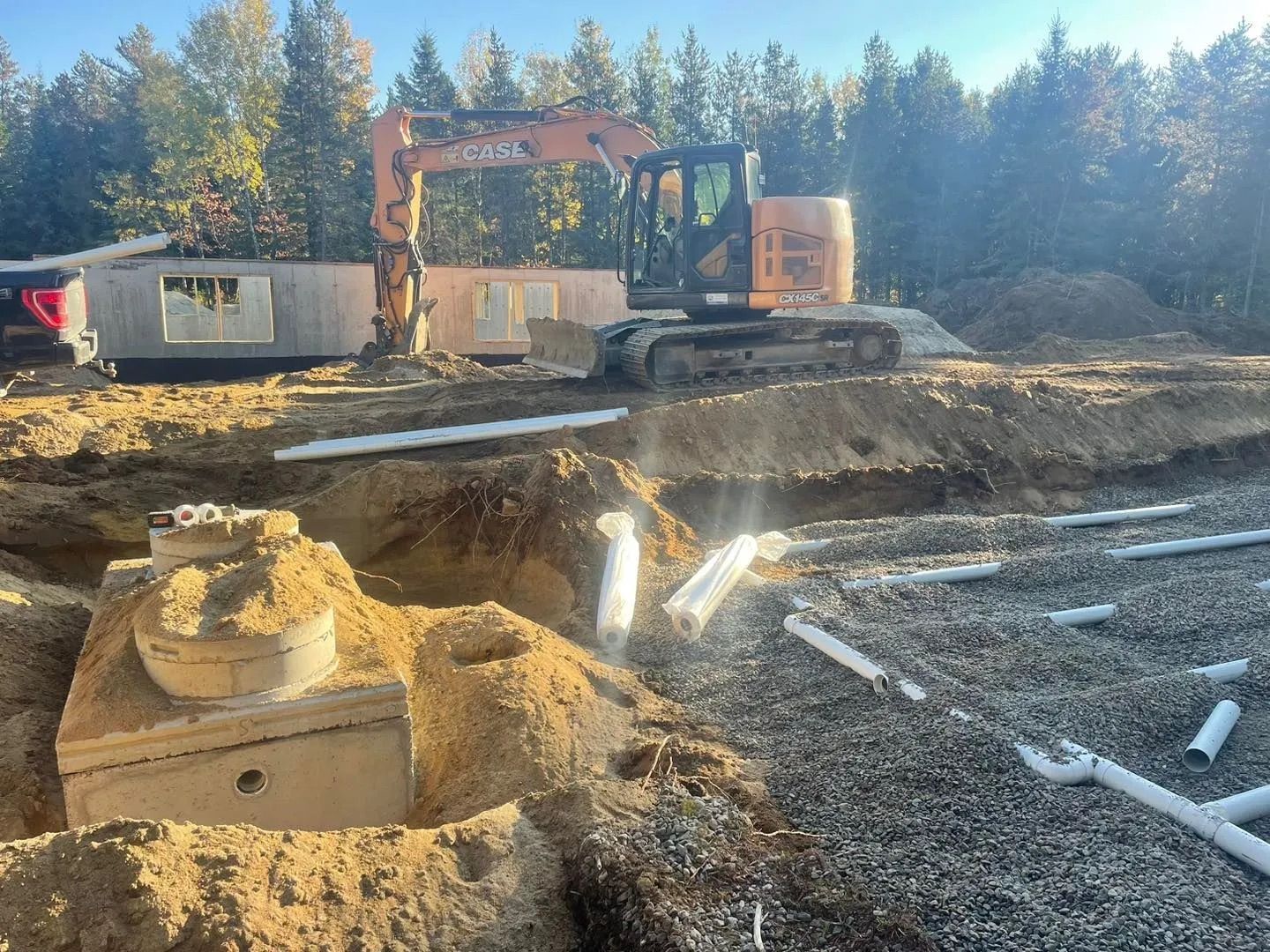 Une pelleteuse orange stationnée sur un chantier de construction, avec des éléments de fosse septique apparents et un sol recouvert de gravier.
