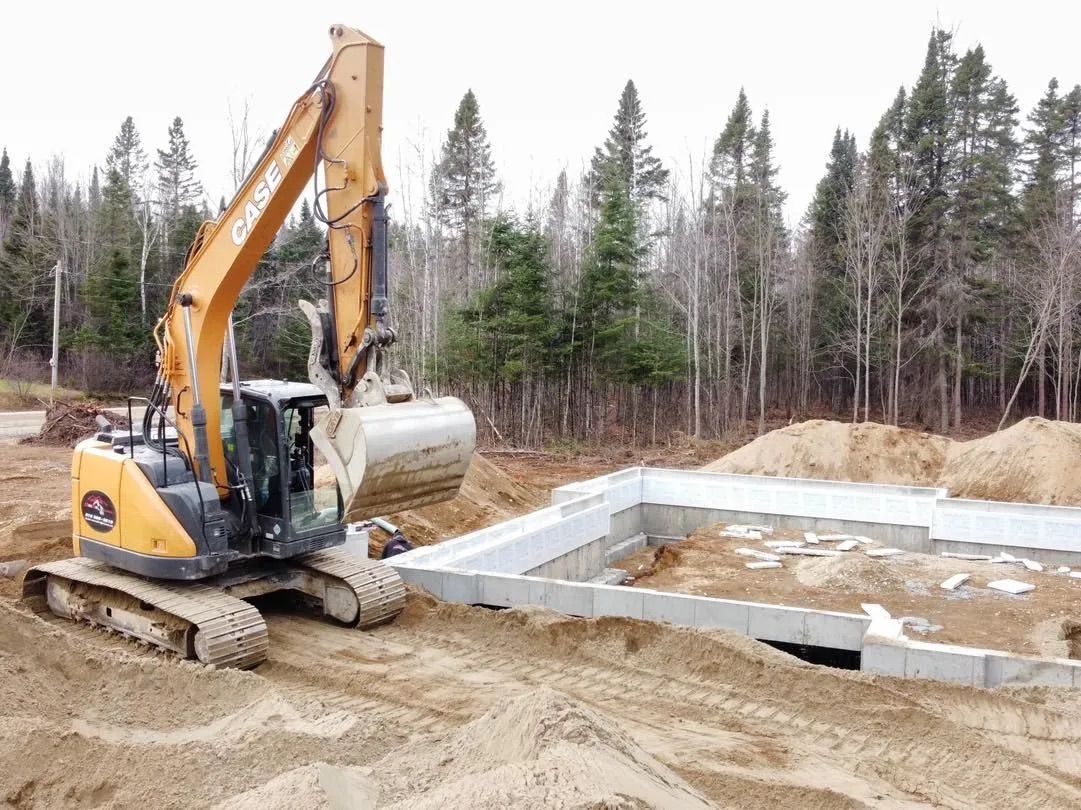 Une pelleteuse jaune descend une grande fosse septique cylindrique dans une tranchée jouxtant les fondations en béton d'une maison.