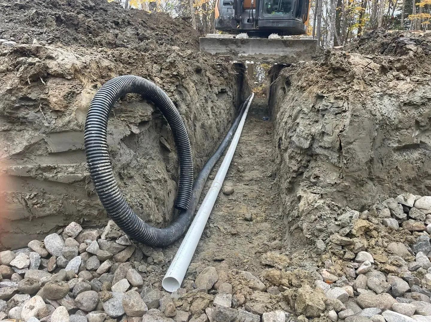 Un tuyau ondulé noir et un tuyau en PVC blanc gisent dans une tranchée boueuse remplie de gravier, avec des engins de chantier visibles.