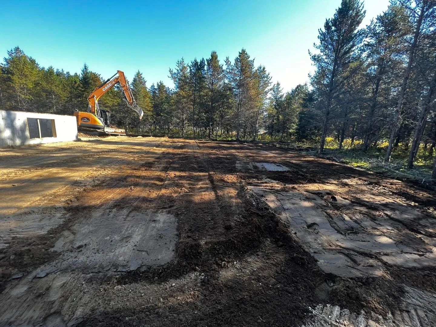Une pelleteuse jaune travaille sur un terrain défriché et boueux, adjacent à une structure blanche et à une forêt, sous un ciel bleu limpide.