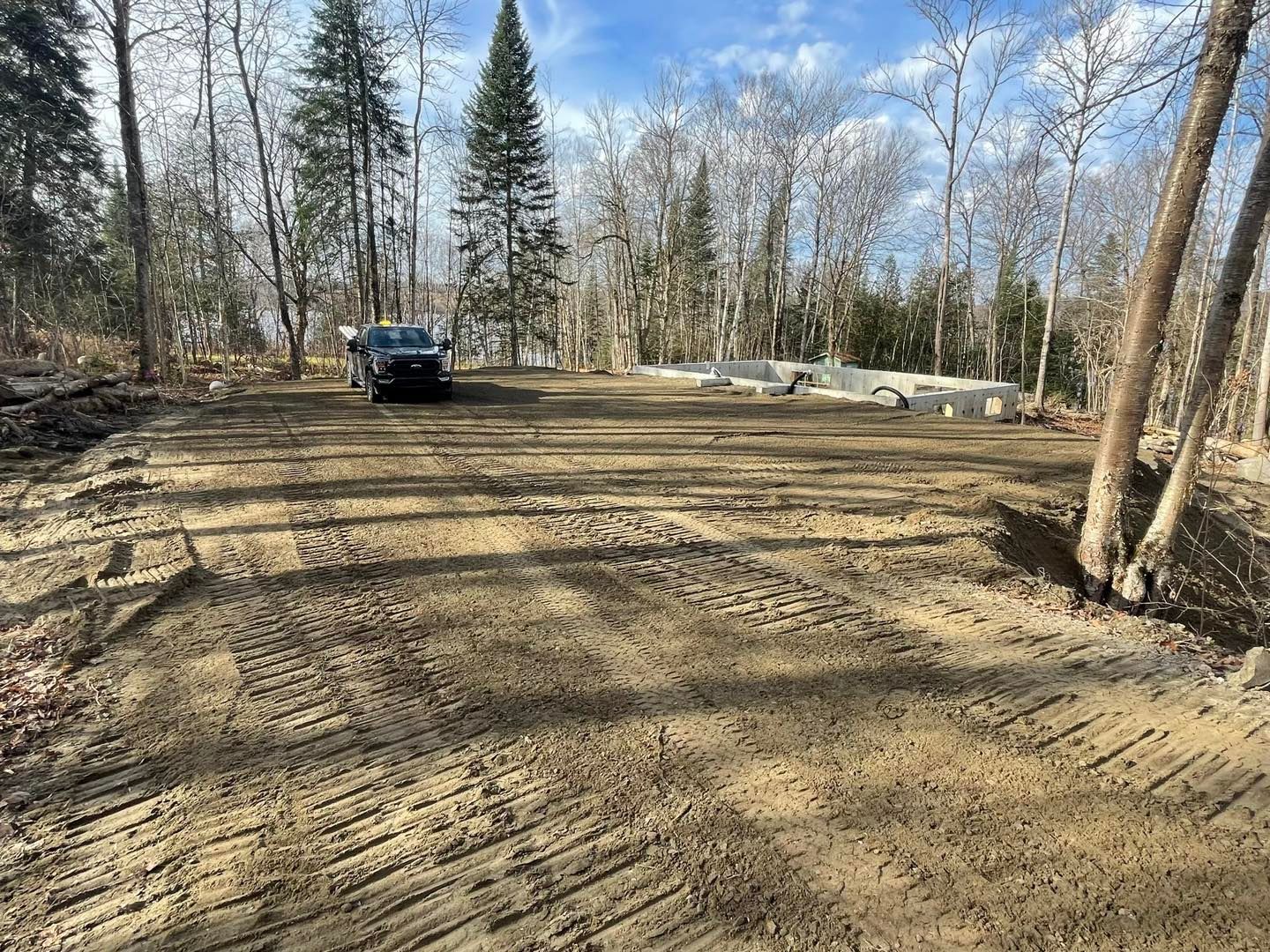 Un camion sombre stationné sur un terrain vague boueux et dégagé, avec des traces de pneus visibles, entouré d'arbres et d'une dalle de béton.