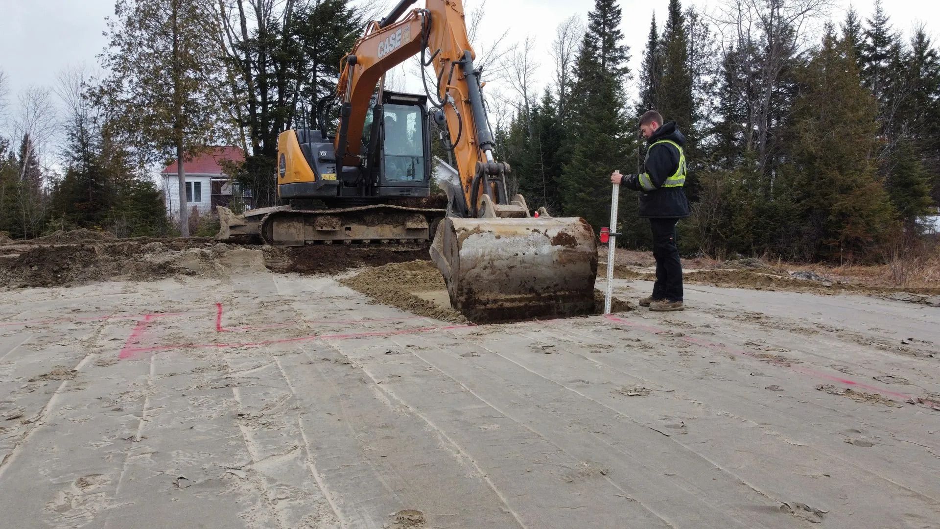 Un conducteur d'excavatrice utilise une mire pour vérifier la profondeur d'un chantier de construction marqué à la peinture rose.