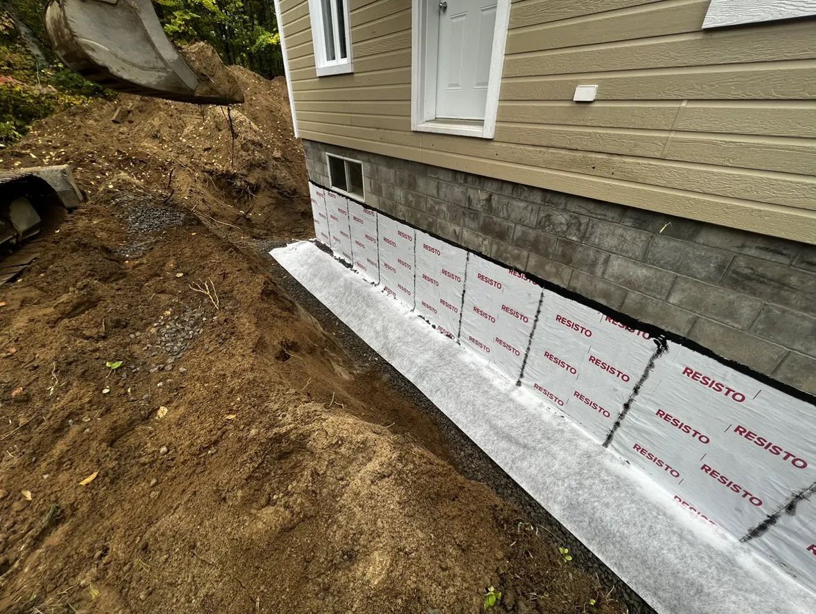 Les fondations d'une maison beige, partiellement excavées, laissent apparaître des rangées de panneaux isolants blancs fixés aux blocs de béton.