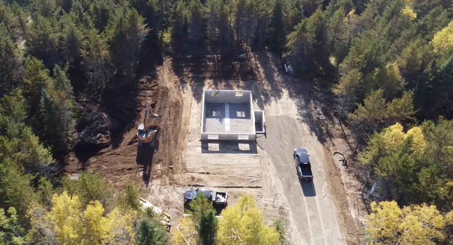 Vue aérienne des fondations d'un bâtiment rectangulaire en construction dans une forêt, avec deux véhicules stationnés à proximité.