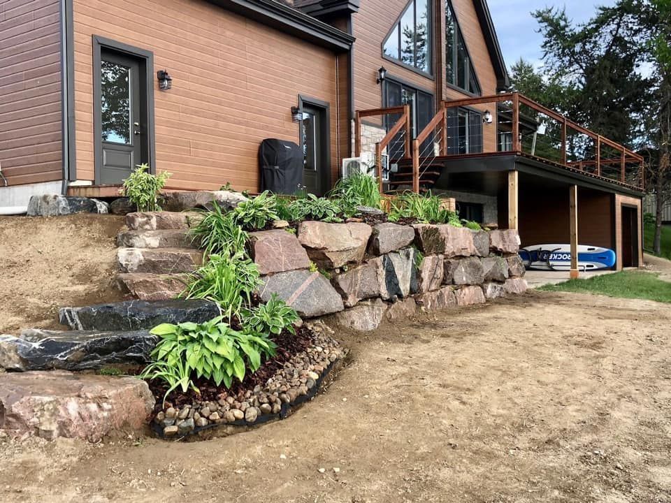 Un grand mur de soutènement en pierre et des marches en pierre mènent à l'entrée latérale d'une maison moderne brune avec une terrasse.