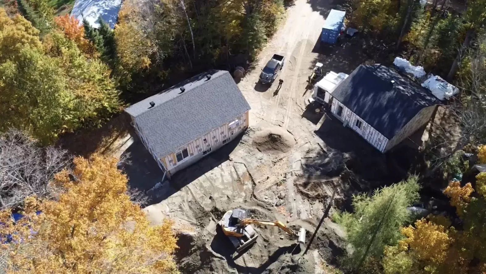 Vue aérienne d'un chantier de construction montrant deux charpentes de maisons, un camion stationné et une excavatrice travaillant sur un terrain en terre.