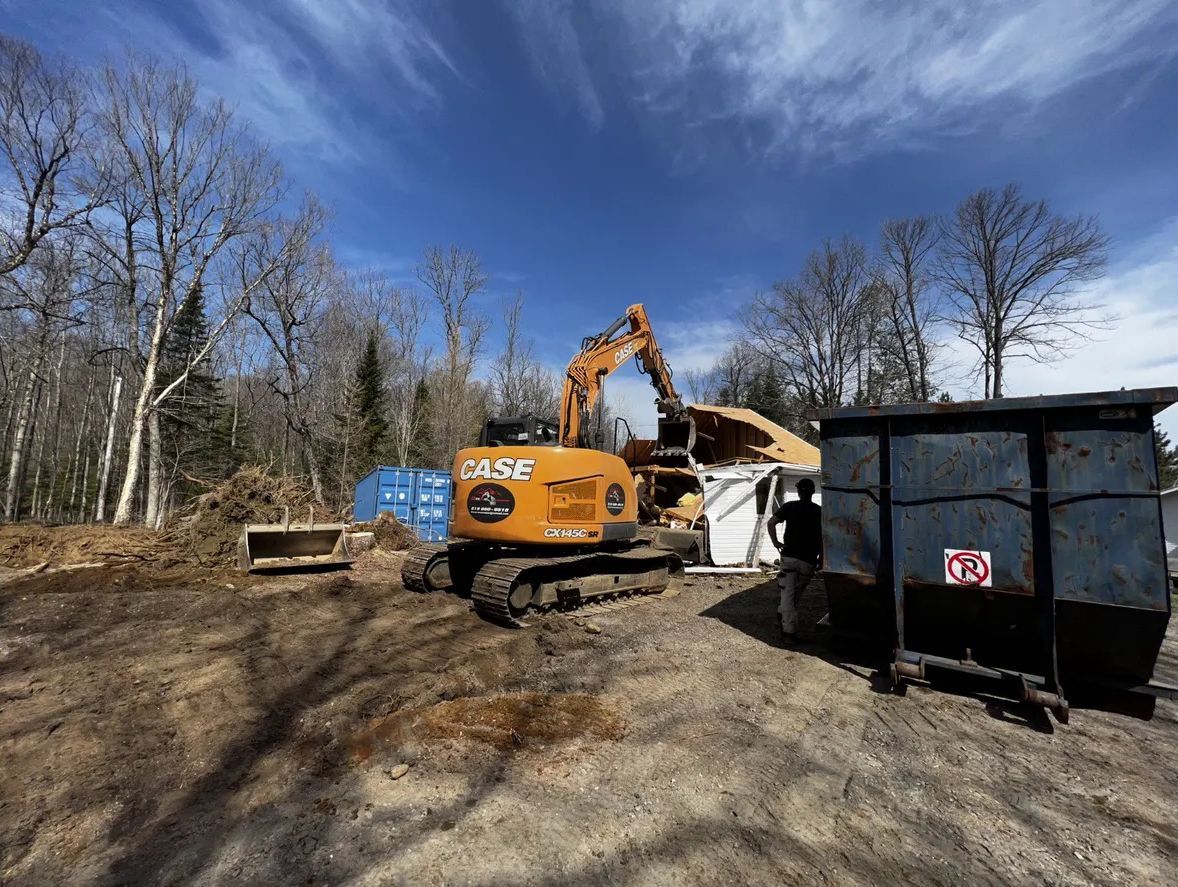 Une pelleteuse jaune déplace des débris sur un chantier de démolition sous un ciel bleu éclatant, avec une benne à ordures au premier plan.