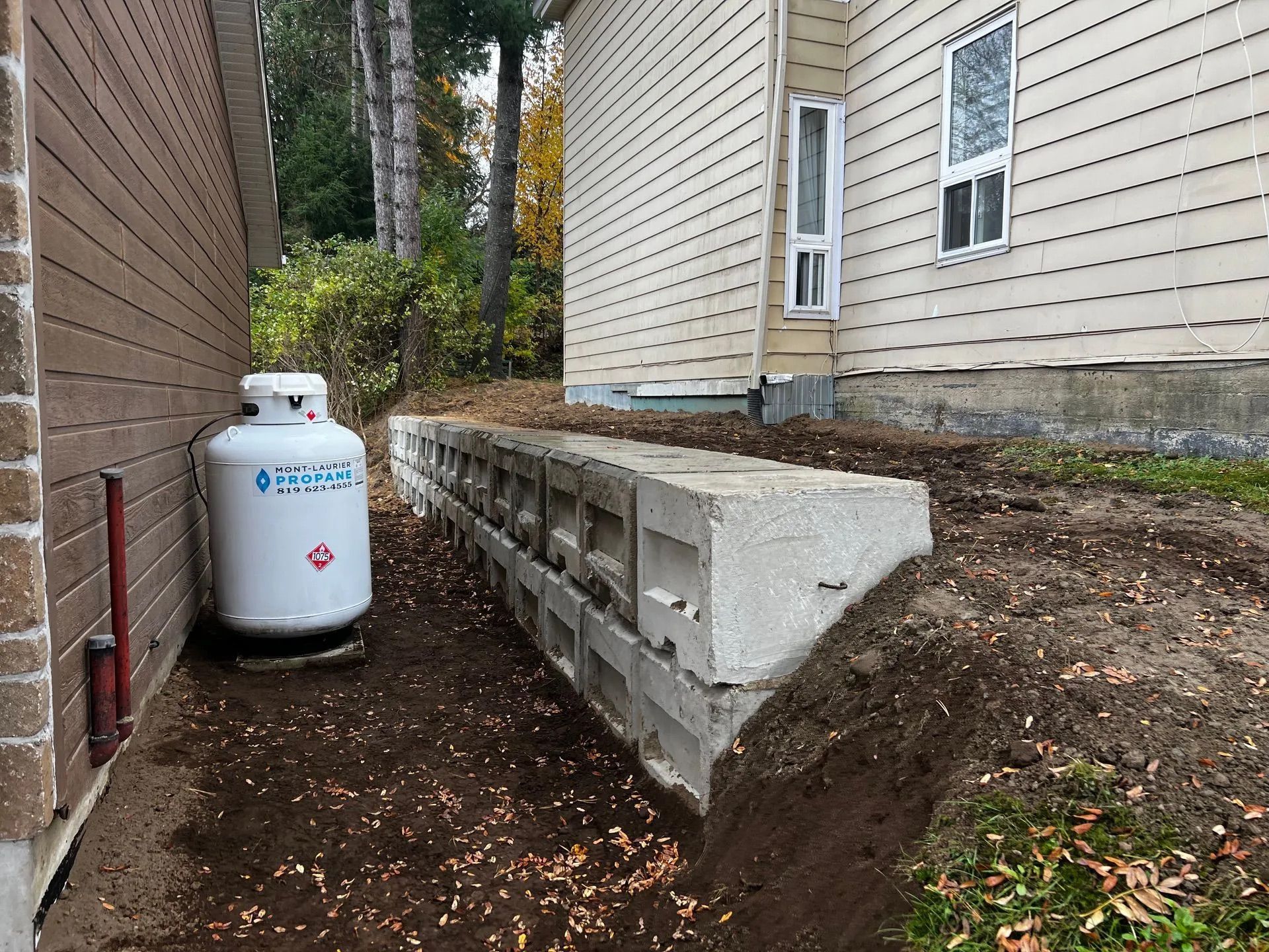 Une bonbonne de propane blanche est posée dans un espace terreux entre un mur de maison brun et un mur de soutènement en blocs de béton à plusieurs niveaux.