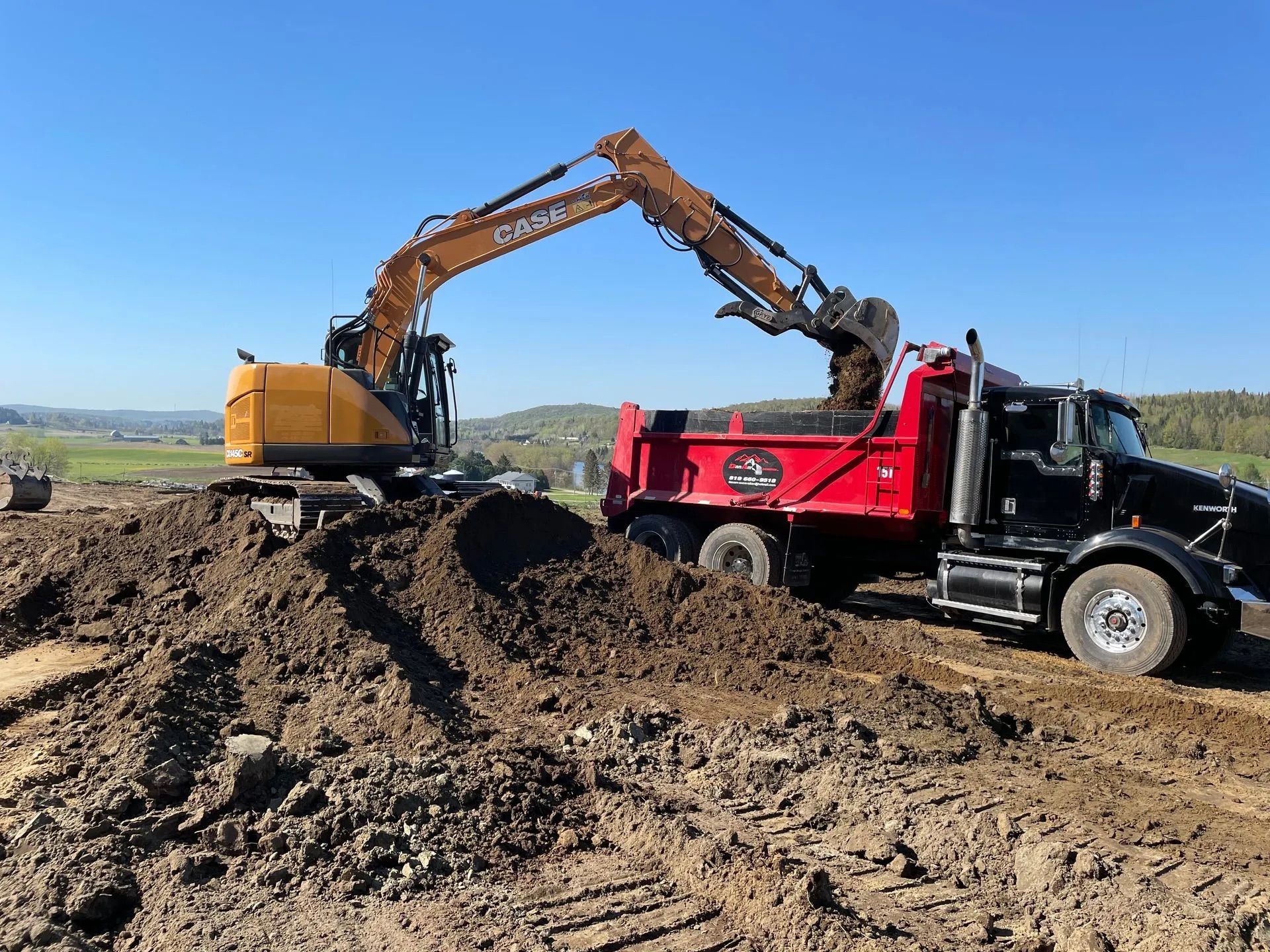 Sur un chantier de construction, sous un ciel dégagé, une pelleteuse jaune charge de la terre sombre à l'arrière d'un camion-benne rouge et noir.