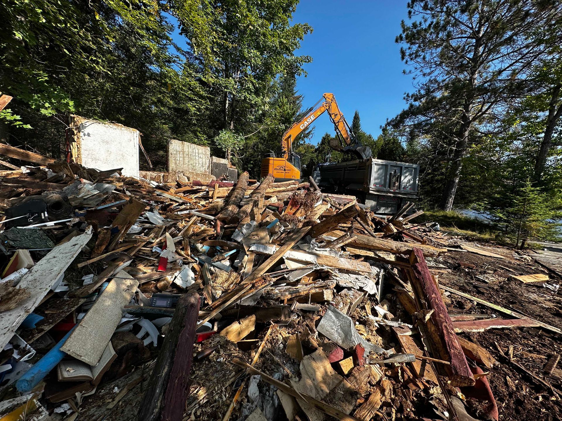 Une excavatrice travaille à dégager un important tas de débris de bois et de gravats de construction près d'une zone boisée, sous un ciel bleu.