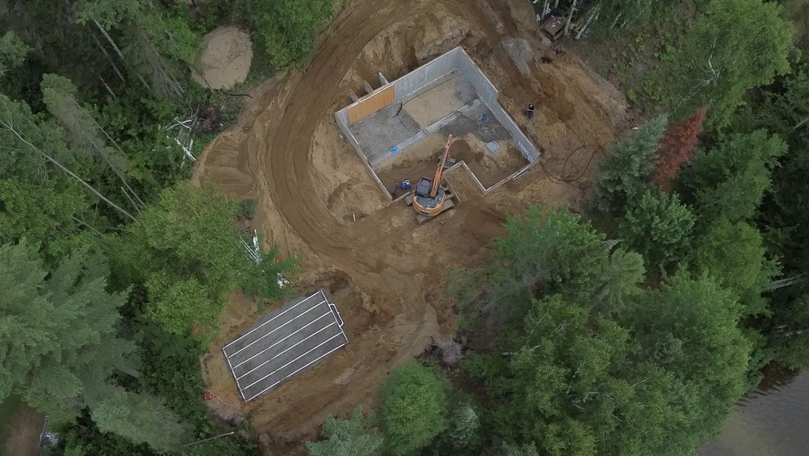 Vue aérienne d'un chantier de construction boisé avec des fondations excavées et un champ d'épandage septique en cours d'installation.
