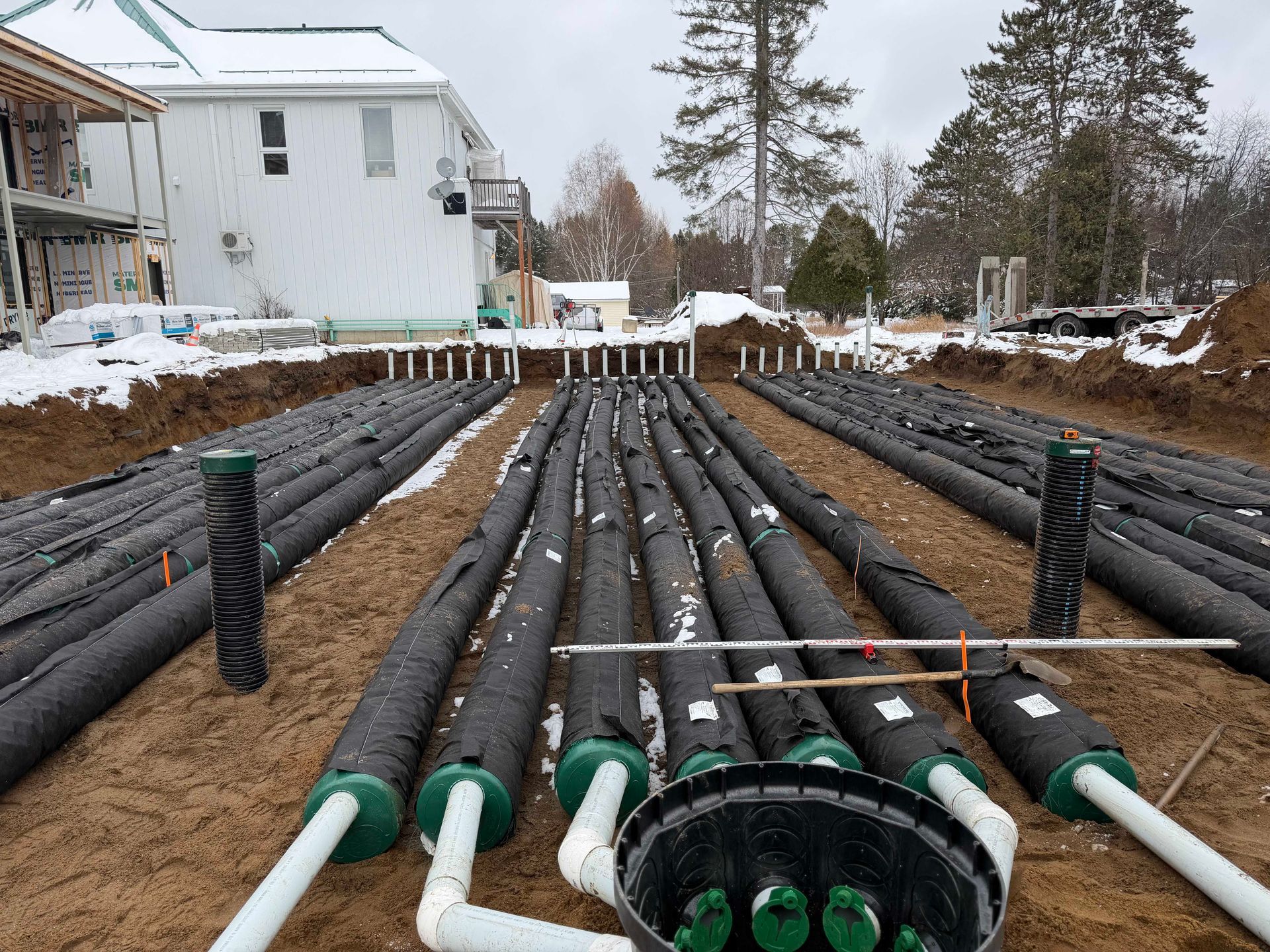 Des rangées de tuyaux de drainage noirs, recouverts de tissu, sont installées dans une tranchée sur un chantier de construction enneigé.